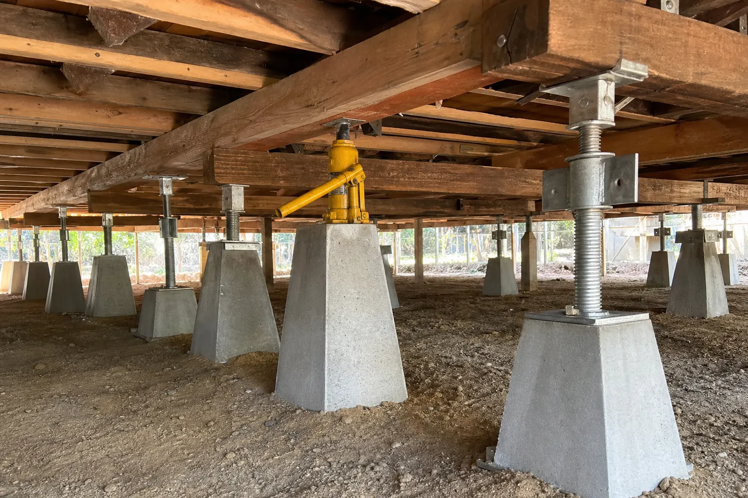 New galvanised steel and concrete stumps supporting the timber bearers under a raised house, with a hydraulic jack mid-lift